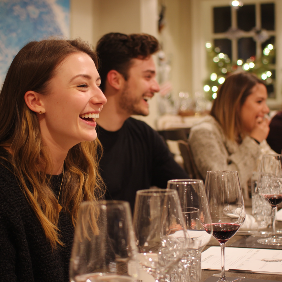 Group of friends laughing at a dinner table with wine glasses and festive decor.