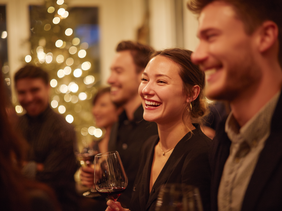 Group of smiling friends enjoying a festive gathering with drinks and warm, twinkling lights.